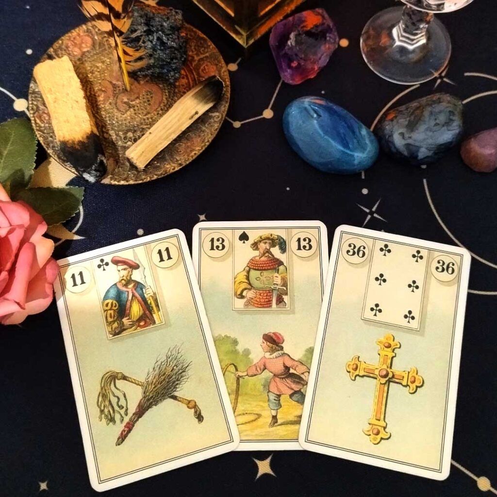 Foto com incenso de palo santo, pedras, cristais e uma rosa na altura do centro da foto à esquerda. Na parte inferior, três cartas do baralho Lenormand: 11 - O chicote; 13 - A criança; 36 - A cruz.