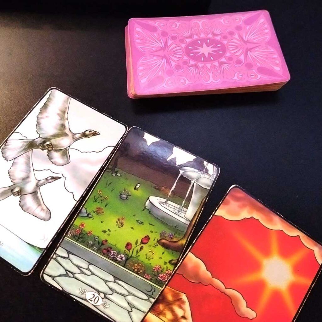 Foto de um baralho cigano com o verso das cartas em tons de magenta. Na parte superior, a pilha de cartas e abaixo, três cartas dispostas em diagonal descendente da esquerda para direita, com as cartas: Os pássaros, O Jardim e O Sol. Ilustrações de cores vibrantes, vívidas. As Leituras de cartomancia são uma das formas de oráculo.