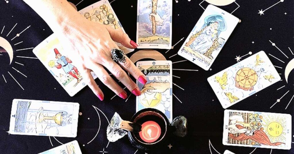 Foto de tiragem de tarô com várias cartas em círculo e uma ao centro. A mão esquerda da Sacerdotisa de Oráculos Oraculares sobrepõe do lado esquerdo da foto duas cartas, com um anel de obsidiana no dedo indicador, unhas bem feitas com esmalte magenta.