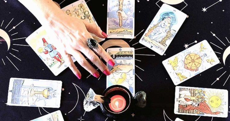 Foto de tiragem de tarô com várias cartas em círculo e uma ao centro. A mão esquerda da Sacerdotisa de Oráculos Oraculares sobrepõe do lado esquerdo da foto duas cartas, com um anel de obsidiana no dedo indicador, unhas bem feitas com esmalte magenta.